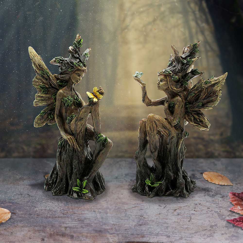 Two carved wooden fairy statues with leafy wings facing each other on a rustic surface with autumn leaves nearby, as if in conversation.