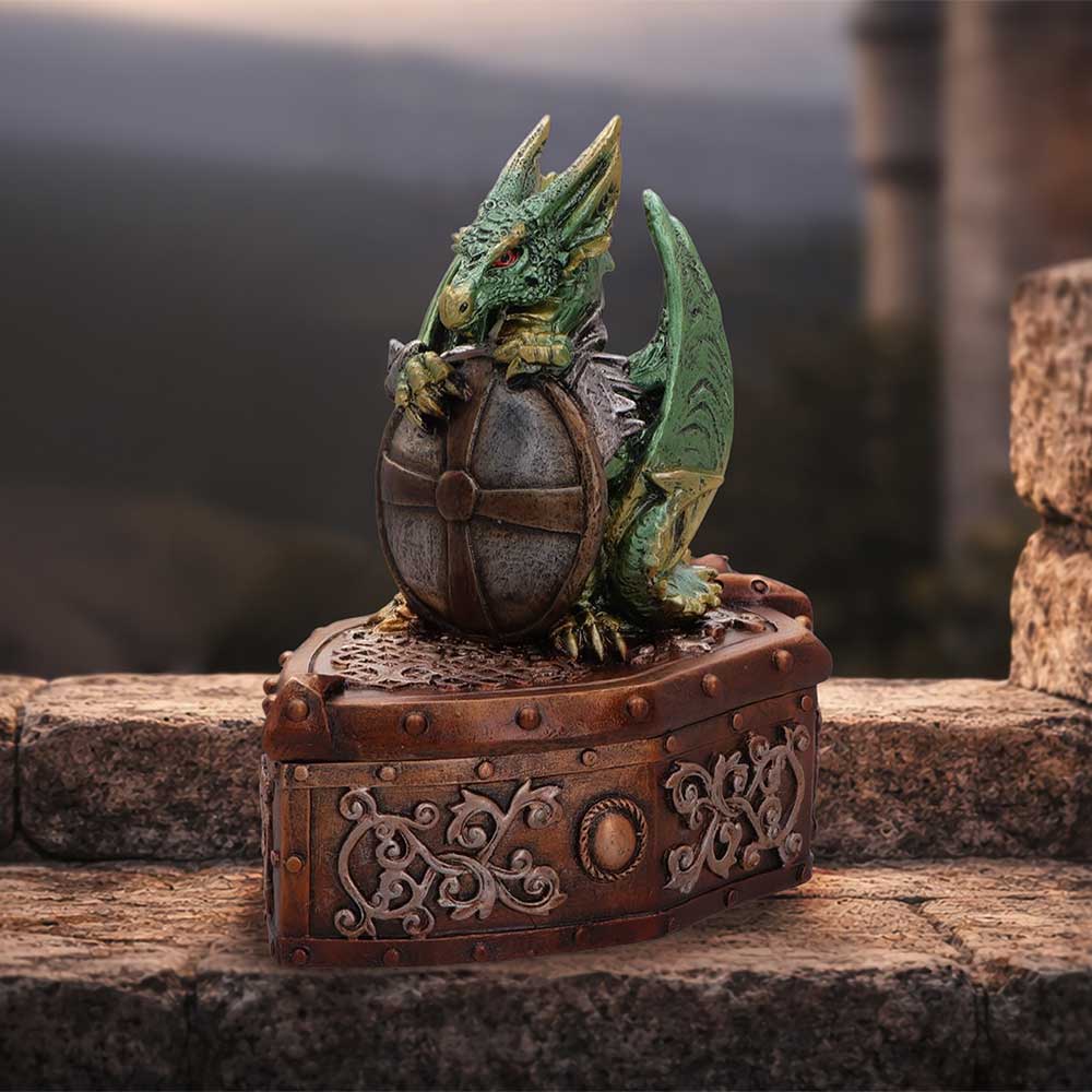 Green dragon figurine perched on a bronze treasure chest with intricate metalwork on a stone ledge; blurred landscape in the background.